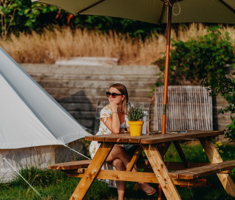 Vrouw aan picknicktafel onder parasol bij REBL Outdoor Bell tent glamping in Friesland, Nederland.