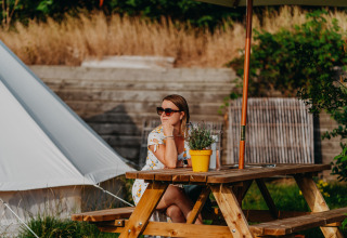 Femme assise à une table de pique-nique sous un parasol au glamping REBL Outdoor Bell tent à Friesland, Pays-Bas.