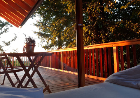 Zicht vanuit bed in Sankt Martin boomhut op houten terras met tafel, stoelen en bomen eromheen.