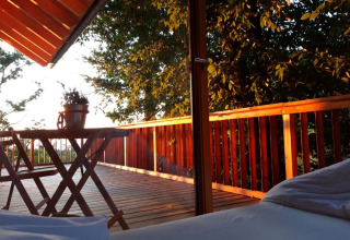 Uitzicht vanuit bed in Sankt Martin boomhut over houten terras met tafel en stoelen, omgeven door bomen.