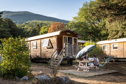 To personer nyder udendørs spisning ved en træhytte på Huttopia Divonne Les Bains glampingplads i Rhône-Alpes.