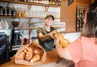 Une vendeuse souriante tend une baguette de pain frais à une cliente dans une boulangerie chaleureuse et accueillante.