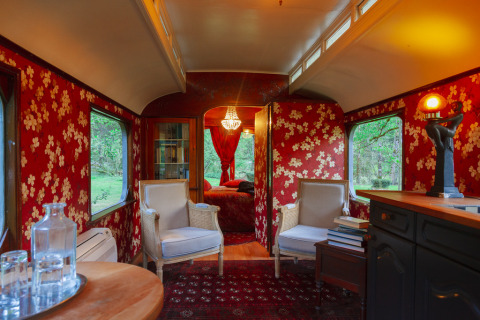 Vintage ingerichte glampingwagon met rode bloemenbehang, fauteuils en ramen met uitzicht op de natuur.
