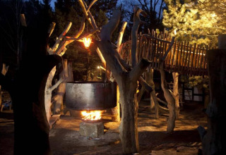 Boomhut-glamping in Saksen bij nacht, grote ketel boven kampvuur, houten brug en sfeervolle verlichting.