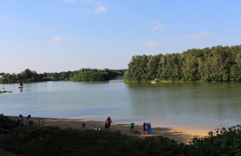 Uitzicht op meer en strand bij Campingpark Heidewald met bomen, zeilboot en mensen op het zand.
