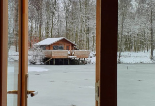 Blick durch Fenster auf eine Holzhütte am zugefrorenen See, umgeben von verschneiten Bäumen in Nord-Ostsee Camp.