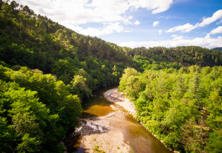 Luftfoto af Camping Les Drouilhedes i Occitanie ved flod, omgivet af frodig grøn skov og campingtelte.