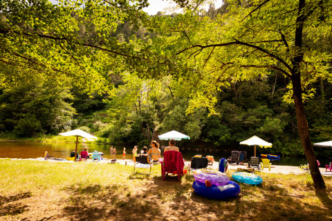 Camping Les Drouilhedes en Occitania: personas disfrutan junto al río, con sombrillas, sillas y flotadores de piscina.