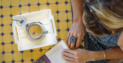 A person reads a book at a tiled table with coffee and a glass at Camping Les Drouilhedes Tent Lodges Occitanië.