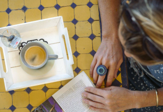 Persona leyendo un libro con café y vaso en una mesa de azulejos en Camping Les Drouilhedes Tent Lodges Occitanië.