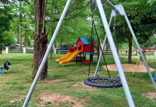 Playground with swings, slides and playhouse surrounded by trees at Camping Les Drouilhedes - Tent Lodges Occitanië.