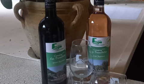 Due bottiglie di vino e due bicchieri con etichette Camping Les Drouilhedes su un tavolo davanti a un vaso.