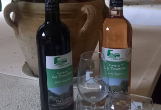 Deux bouteilles de vin et deux verres avec étiquettes Camping Les Drouilhedes sur une table devant un pot.