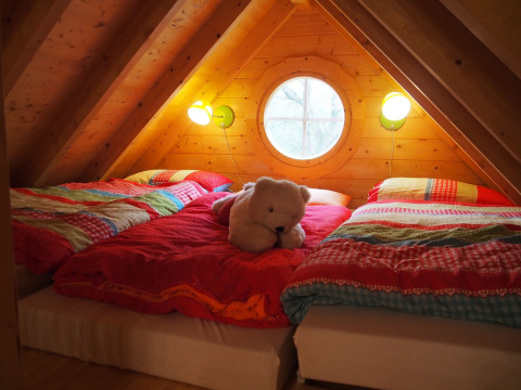 Cozy sleeping area in a treehouse with colorful bedding, a teddy bear, and a round window in the attic.