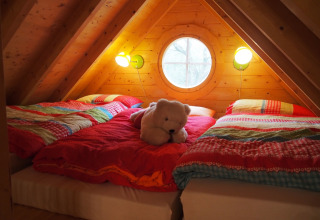 Knusse slaapruimte in een boomhut met kleurrijk beddengoed, knuffelbeer en rond raam op de zolder.