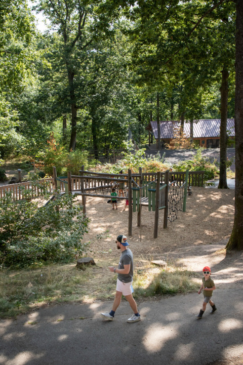 Een gezin wandelt langs een speeltuin in het bos bij Huttopia Wattwiller - Glamping Grand Est.
