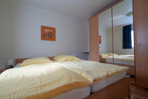 Helles Schlafzimmer mit Doppelbett, gelber Bettwäsche, Wandbild und großem Kleiderschrank mit Spiegel.