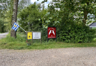 Ingang van Mini Camping De Bokkesprong met vlag, borden, groene bomen en glamping- of chaletverhuur.