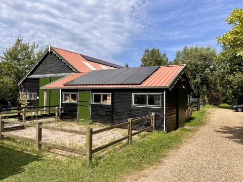 Sort træbygning med solpaneler på taget ved Glamping De Bokkesprong, omgivet af træer og grusvej.