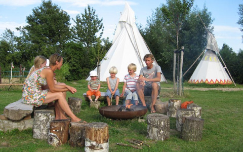 Familie nyder lejrbål foran tipi-telte på Glamping - De Bokkesprong, omgivet af grøn natur.