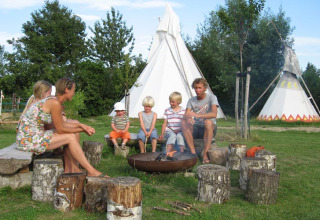 Familie nyder lejrbål foran tipi-telte på Glamping - De Bokkesprong, omgivet af grøn natur.