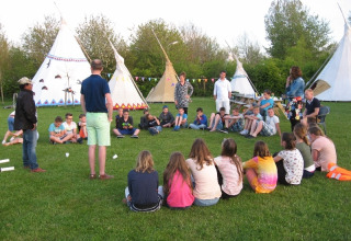 Kinderen en volwassenen zitten in een kring voor tipi’s bij Glamping - De Bokkesprong tijdens buitenactiviteiten.