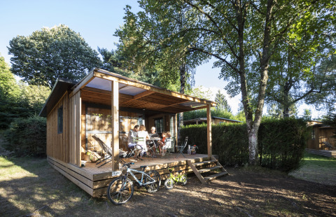Gezin ontspant op de veranda van een houten hut bij Huttopia Lac de la Siauve - Glamping Auvergne, omringd door bomen.