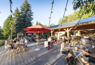 People dining outdoors on a wooden terrace surrounded by trees at Huttopia Lac de la Siauve Glamping site.