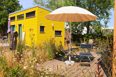 Tiny lodge - Ellertshäuser See, gelbe Glamping-Unterkunft mit Sonnenschirm, Gartenmöbeln und Blumen davor.