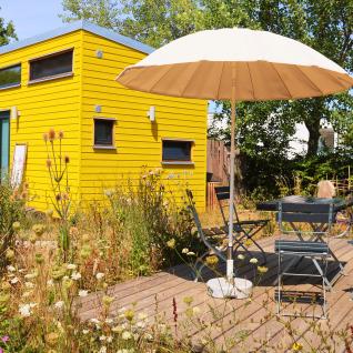 Tiny lodge - Ellertshäuser See, yellow glamping accommodation with patio umbrella and seating in a flowery garden.