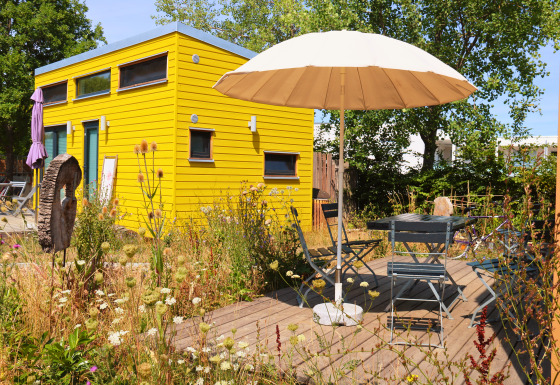 Tiny lodge - Ellertshäuser See, gele glamping accommodatie met parasol en zitje in een bloemenrijke tuin.