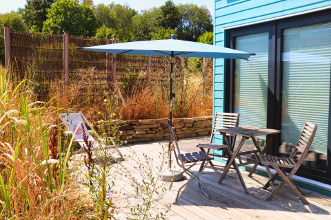 Buitenterras bij Tiny lodge - Ellertshäuser See met houten meubels, blauwe parasol en wilde begroeiing.