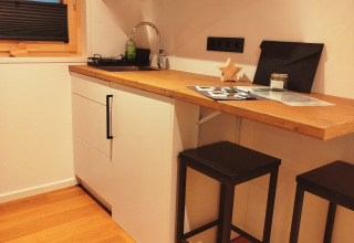 Moderne tiny lodge keuken met houten blad, zwarte barkrukken, witte kasten en raam met uitzicht op lucht.