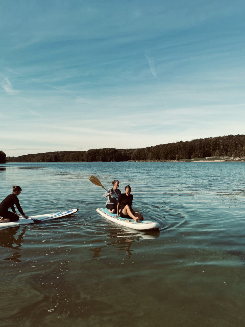 To personer på paddleboard og én ved søen, under blå himmel nær Tiny lodge - Ellertshäuser See glamping.
