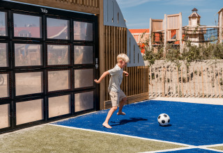 Niño juega al fútbol descalzo en área de juegos moderna de Strandpark De Zeeuwse Kust - Safaritenten Zeeland.