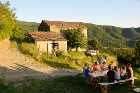 Grupo de personas comiendo al aire libre en una mesa larga cerca de antiguas construcciones de piedra y colinas.