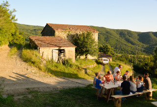 Group of people dining outdoors at a long wooden table by rustic stone buildings in a scenic hilly area.