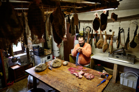 Hombre corta carne fresca en una mesa de madera, rodeado de embutidos colgados en Novanta Skylodge, Italia.