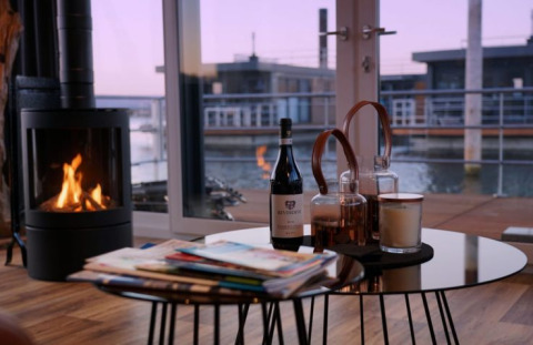 Escena interior de Floating Village Brombachsee - Lodges Bayern con chimenea, vino y vista al agua.