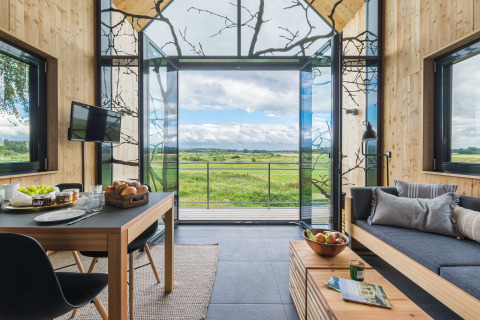 Moderne glampingindkvartering med store vinduer, udsigt over grønne marker og hyggelig indretning.