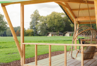 Glamping ved Landrijk de Reesprong med hyggelig træterrasse og hængestol, udsigt til grøn mark og telte.