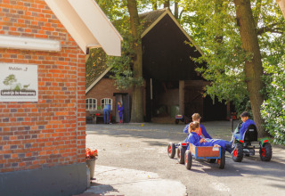 Børn leger udenfor på gokarts ved Glamping - Landrijk de Reesprong, omgivet af træer og bondebygning.