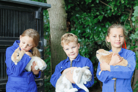 Trois enfants en combinaisons bleues tiennent des lapins à 'Glamping - Landrijk de Reesprong', entourés de verdure.