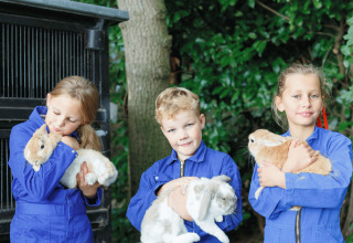 Drei Kinder in blauen Overalls halten Kaninchen bei 'Glamping - Landrijk de Reesprong' in der Natur.