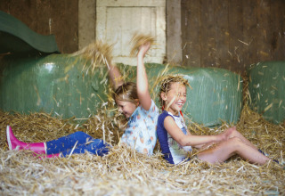 Two happy children play in straw at Glamping - Landrijk de Reesprong, surrounded by hay bales in a barn.