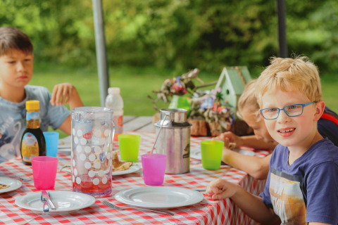 Bambini che gustano un pasto all'aperto a Glamping - Landrijk de Reesprong, seduti a un tavolo con tovaglia rossa.