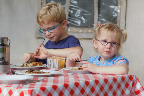 Two children with glasses enjoy pancakes at a red-checkered table at Glamping - Landrijk de Reesprong.