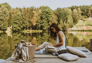 Woman relaxing by lakeside dock with pillows and basket at Falkensteiner Premium Camping Blaguš, Slovenia.