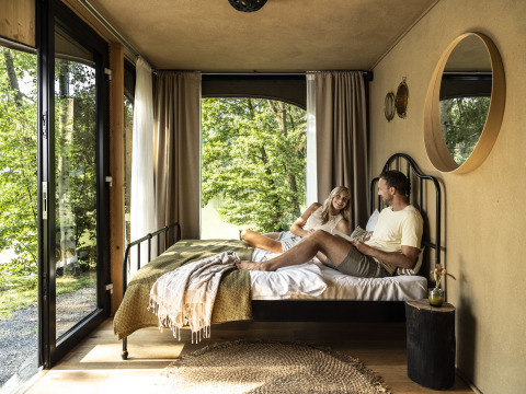 A couple relaxes in a bright, modern glamping cabin with forest views at Falkensteiner Premium Camping Blaguš in Slovenia.