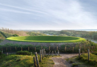Grassy hill with a large circular dip, surrounded by pathways and fences at HaagseStrandhuisjes in The Hague.